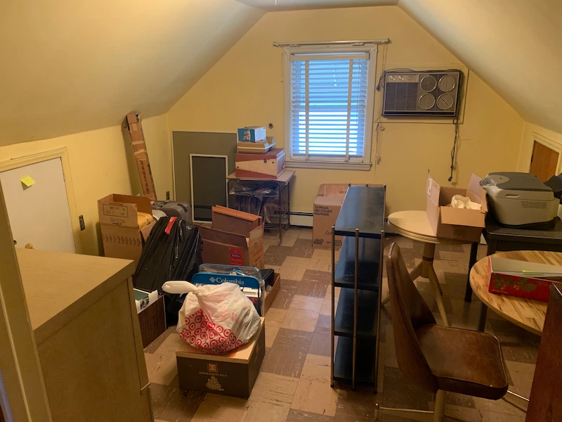 An attic storage room in Glen Ridge, North Jersey filled with a large pile of old cardboard boxes, miscellaneous furniture, and a window A/C unit before being cleared by JunkDoctors.