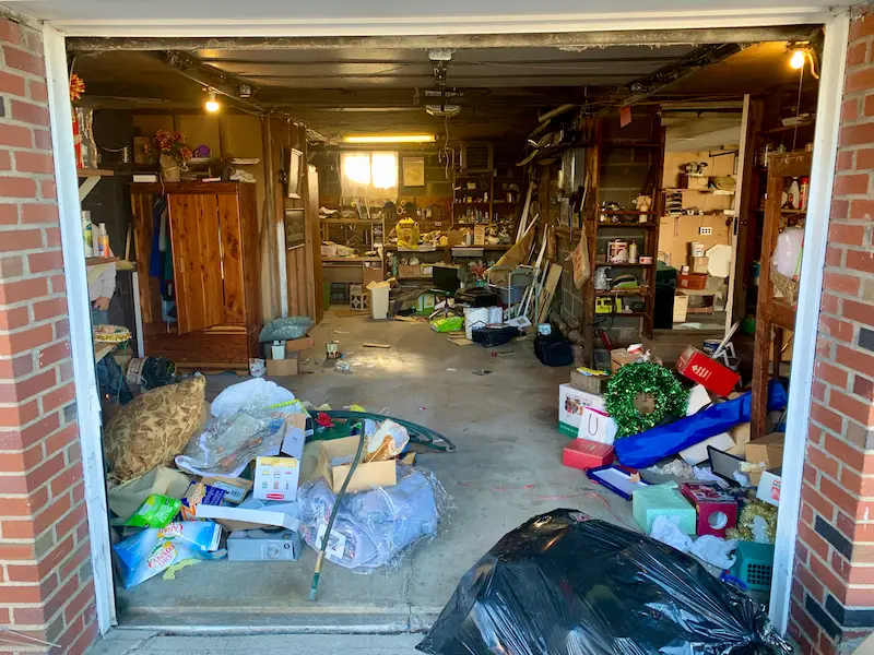 A messy garage filled with household junk, Christmas decor, tools, boxes, and old furniture before a Junk Doctors cleanout.