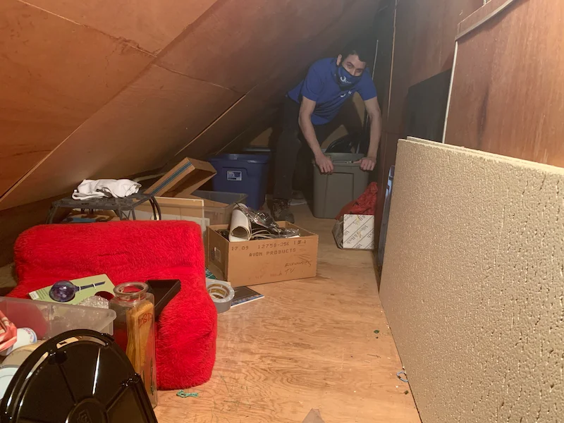A heavily cluttered New Jersey attic space with exposed wooden beams, filled with cardboard boxes, old furniture, and household junk before removal.