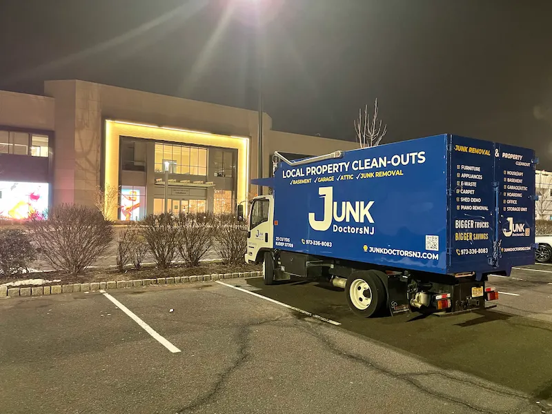 A large blue Junk Doctors NJ property clean-out truck parked in a commercial parking lot outside a brightly lit modern retail building at night.