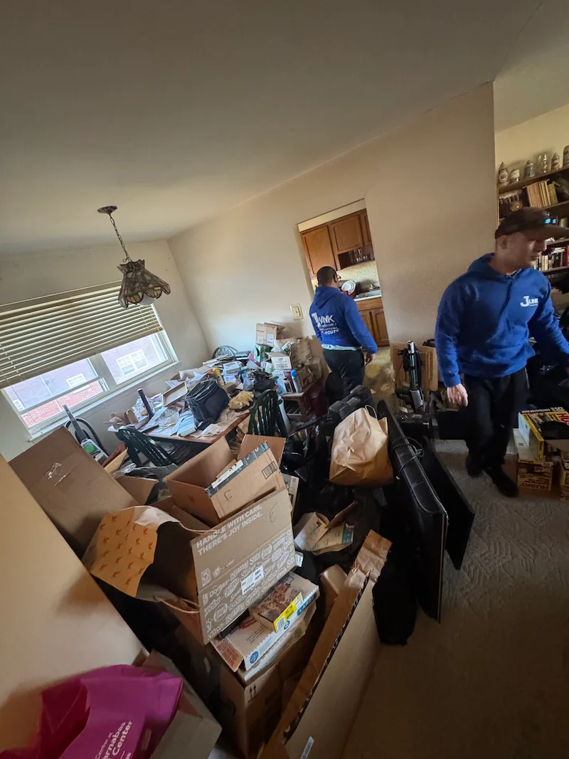 Before photo showing an extremely cluttered apartment room packed floor-to-ceiling with cardboard boxes, bags, and loose debris requiring a full JunkDoctors cleanout in NJ.